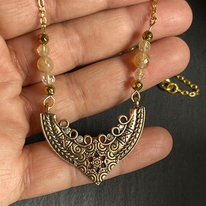 May include: Gold necklace with a crescent moon pendant featuring intricate details and small clear stones. The necklace is made with gold chain and beads.