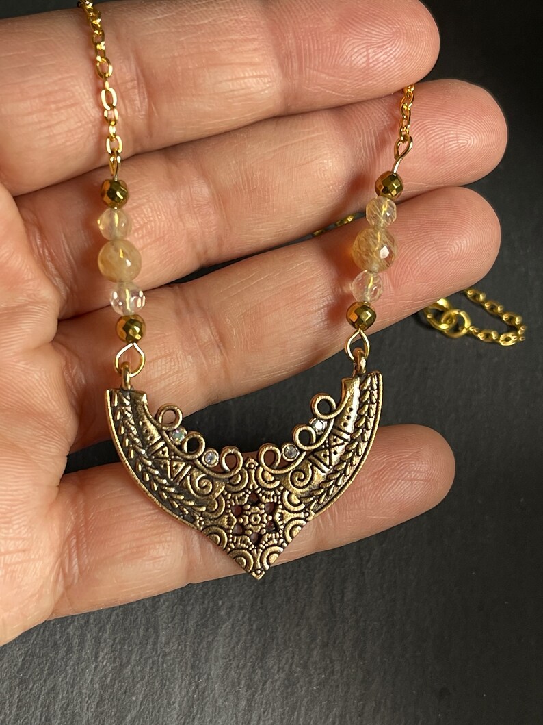 May include: Gold necklace with a crescent moon pendant featuring intricate details and small clear stones. The necklace is made with gold chain and beads.