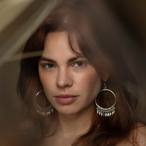 May include: A woman with long, auburn hair wearing large silver hoop earrings with dangling charms. The earrings have a hammered metal finish.