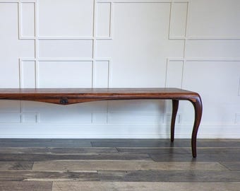 May include: A long, dark brown wooden coffee table with curved legs. The table has a slightly curved top and a small decorative element in the center. The table is set against a white wall with a geometric pattern.