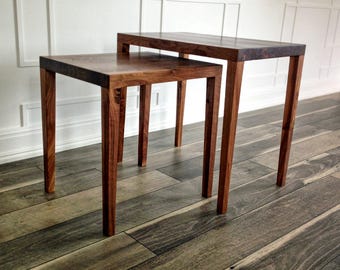 May include: Two wooden nesting tables with a dark brown finish. The tables have square tops and four slender legs. The larger table is 24 inches wide, 24 inches deep, and 20 inches tall. The smaller table is 18 inches wide, 18 inches deep, and 16 inches tall.