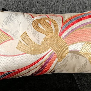 May include: Rectangular decorative pillow with a cream-colored background and a large gold embroidered bow design. The pillow features red, pink, purple, and blue accents. The background has a subtle pattern of cranes.