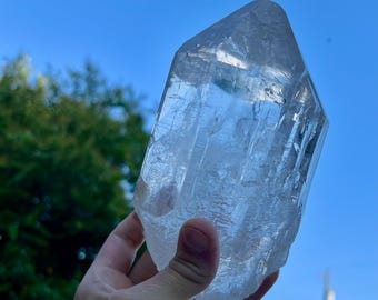 May include: A large, clear quartz crystal with a pointed tip. The crystal is held in a hand against a blue sky background.