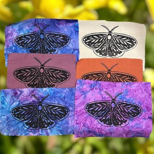 May include: A collection of fabric squares in various colors, including blue, purple, and orange. Each square features a black moth design. The fabrics have a tie-dye or batik appearance, with the moth design prominently displayed.