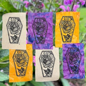 May include: A collection of six fabric patches with a black floral design inside a coffin shape. The patches are in various colors, including beige, purple, and orange, with a floral design.