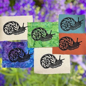 May include: A collection of six fabric squares, each featuring a black snail design with a leafy pattern. The squares are in various colors, including beige, green, blue, orange, and purple, set against a blurred floral background.