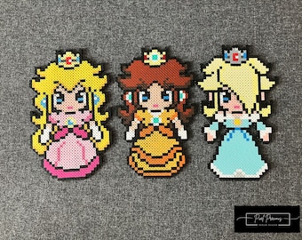 May include: Three pixel art figures of princesses from the Super Mario Bros. video game series. Peach is wearing a pink dress with a crown, Daisy is wearing a yellow dress with a crown, and Rosalina is wearing a blue dress with a crown.