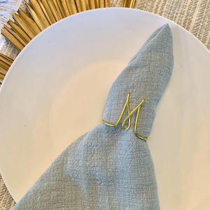 May include: A gold metal napkin ring shaped like the letter "M" holding a light blue napkin on a white plate.