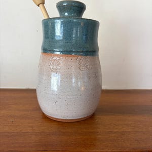 May include: A handmade ceramic honey pot with a teal blue glazed top and lid, and a light beige lower section with embossed floral designs. A wooden honey dipper rests inside. The pot sits on a wooden surface.
