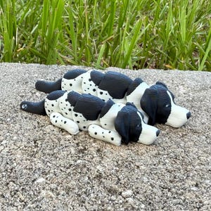 Puede incluir: Dos figuras decorativas de perros en posición acostada. Los perros son negros y blancos, con manchas negras en sus cuerpos. Los perros tienen orejas negras y caras blancas. Las figuras descansan sobre una superficie gris.