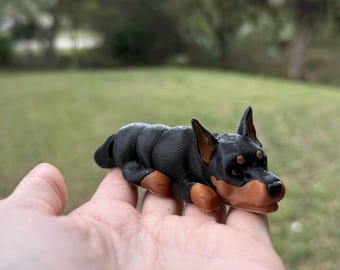 May include: A black and brown 3D printed figurine of a dog lying down. The dog has a short, smooth coat and is posed in a sleeping position.