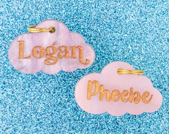 Può includere: Due targhette identificative per animali domestici a forma di nuvola rosa con lettere dorate. La targhetta superiore recita "Logan" e la targhetta inferiore recita "Phoebe".