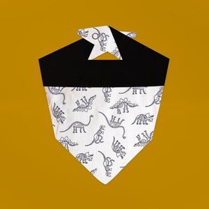 May include: A white bandana with a black border and a pattern of dinosaur skeletons. The bandana is folded into a triangle shape.