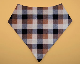 May include: A plaid bandana in a triangular shape, featuring a pattern of black, white, and brown squares. The bandana is set against a solid, light orange background.