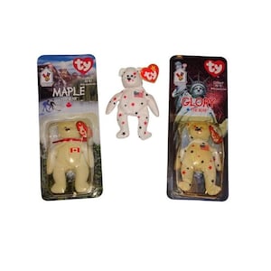 May include: Three Ty Beanie Baby bears. One is white with red stars and an American flag. Another is cream-colored with a red bow and a Canadian flag. The third is cream-colored with red and gold stars and an American flag.