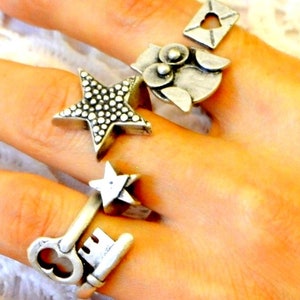 Puede incluir: Una mano muestra varios anillos plateados. Los anillos presentan varios diseños, incluyendo una estrella con detalles punteados, un búho, una llave, una estrella y un sobre con un recorte de corazón. Los anillos probablemente son de metal y tienen una estética vintage o artesanal.