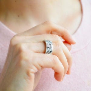 May include: A silver ring with a geometric design is worn on a finger. The ring features a series of parallel lines and rectangular shapes. The ring is made of a shiny metal and has a modern, minimalist style.