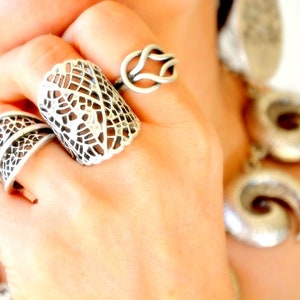 Könnte beinhalten: Nahaufnahme von silbernen Ringen an einer Hand. Die Ringe umfassen ein breites, offenes Design, einen knotenförmigen Ring und einen Ring mit einem gebogenen, gemusterten Band. Im Hintergrund ist ein passender silberner Ohrring zu sehen.