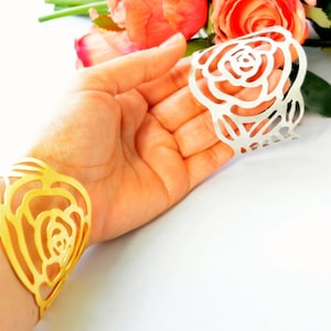 May include: A gold cuff bracelet and a silver ring, both with a rose design. The bracelet is on a wrist, and the ring is held in a hand. The jewellery is set against a white background with pink flowers.
