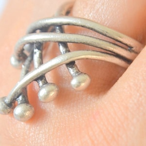 May include: A silver ring with an abstract design. The ring has multiple curved bands that intertwine, with small, spherical accents at the ends of some of the bands. The ring is worn on a finger.
