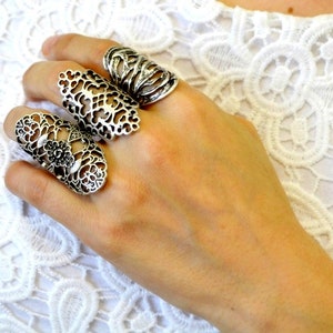 May include: Three silver-toned rings worn on the fingers. The ring on the index finger features an intricate, openwork filigree pattern. The middle finger ring displays a similar ornate, lace-like design. The ring finger ring has a textured, organic, layered metal structure.