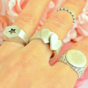Puede incluir: Una mano con varios anillos plateados. Un anillo presenta una estrella, otro dos corazones, un tercero una banda texturizada y el último un óvalo en blanco. Los anillos son todos de color plateado mate.