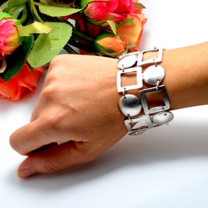 Può includere: Bracciale argentato con forme geometriche, tra cui quadrati, cerchi e ovali. Il bracciale è indossato su un polso, mostrando il suo design e la sua lucentezza metallica. I collegamenti sono connessi, creando un accessorio flessibile ed elegante.