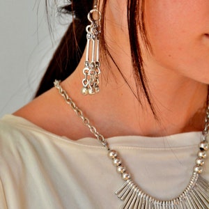 Può includere: Un set composto da una collana e un orecchino pendente color argento. La collana presenta una catena con perline rotonde e un ciondolo centrale a frangia composto da barre metalliche verticali. L'orecchino mostra tre barre con anellini.