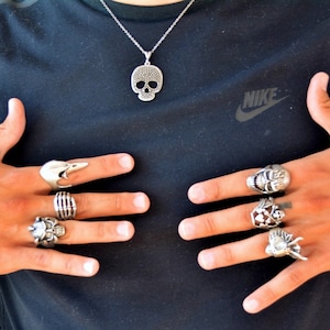 Könnte beinhalten: Silberne Totenkopf-Anhänger-Halskette und mehrere Silberringe. Die Ringe weisen verschiedene Designs auf, darunter Totenköpfe, Skeletthände und abstrakte Formen. Der Schmuck wird vor einem schwarzen Hemd präsentiert.