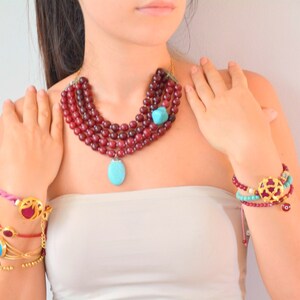 Peut inclure: Un collier multi-rangs composé de perles rouge foncé, d'une pierre turquoise et d'un grand pendentif turquoise. L'ensemble comprend plusieurs bracelets avec des perles rouges et turquoise, des détails en métal doré et des petits charms.