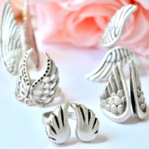 Puede incluir: Una colección de anillos plateados, cada uno con motivos de alas. Los anillos varían en estilo, mostrando diferentes formas y texturas de alas, algunos con detalles intrincados. Los anillos se exhiben sobre una superficie blanca.