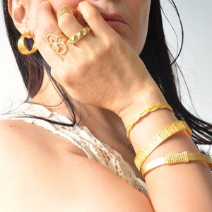 May include: A collection of gold jewelry including hoop earrings, rings, and bracelets. The earrings are multi-layered, and the rings feature various designs. The bracelets include a braided style, a patterned band, and a solid band with a textured clasp.