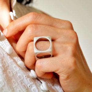 May include: A silver ring with a square design and a circular cutout. The ring is worn on a finger, showcasing its geometric shape and minimalist style. The ring's polished surface reflects light, highlighting its modern aesthetic.