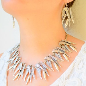 May include: Silver-toned necklace and matching earrings set. The jewelry features a leaf-like design with small, cylindrical accents. The necklace is a choker style, and the earrings hang from a chain. The set is worn against a white background.