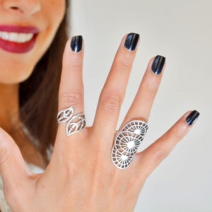 Könnte beinhalten: Eine Hand zeigt zwei silberne Ringe. Ein Ring hat ein Blattdesign, der andere ist ein großer, offener Ring mit einem geometrischen Muster. Die Nägel sind schwarz lackiert. Schmuck.
