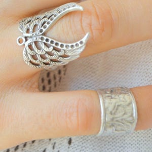 Puede incluir: Se muestran dos anillos de plata. Uno presenta un diseño de ala abierta con detalles de plumas. El otro es una banda lisa con un patrón texturizado. Ambos anillos se llevan en los dedos.