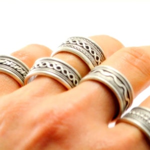 May include: A collection of five silver-toned metal rings worn on fingers. Each ring features a distinct embossed pattern, including braided, wavy, and dotted designs. The bands have a polished finish with darkened recessed areas to highlight the details.