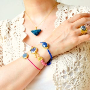 Può includere: Un anello dorato con una pietra blu scuro, una collana a catena dorata con un ciondolo druzy blu triangolare e due braccialetti con pietre blu su cordini rosa e blu con dettagli metallici dorati e una chiusura a gancio.