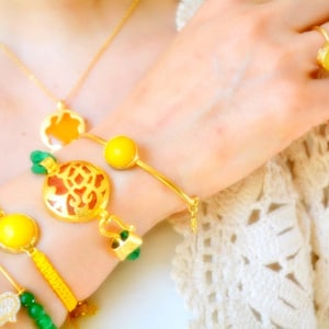 以下が含まれることがあります： イエローの宝石が付いたリングとブレスレットを含む、ゴールドトーンのジュエリーセット。ペンダント付きのゴールドネックレスと、装飾的なチャームが付いたブレスレットも表示されています。ジュエリーは柔らかくニュートラルな背景に配置されています。