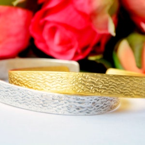 Può includere: Due bracciali aperti, uno color argento e uno color oro, entrambi caratterizzati da una superficie testurizzata con un motivo a onde ripetuto. I bracciali sono sovrapposti, mettendo in risalto la finitura metallica e il design.