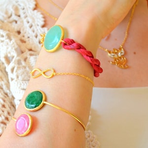 May include: A collection of four bracelets worn on an arm, including a red braided cord bracelet with a mint green oval stone, a gold infinity chain bracelet with small clear stones, and two gold cuff bracelets with green and pink oval stones.