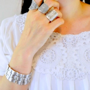 Könnte beinhalten: Eine Auswahl an Silberringen und einem Armband. Die Ringe weisen verschiedene Designs auf, darunter strukturierte Muster und ein rechteckiger Siegelring. Das Armband hat ein Gliederdesign. Der Schmuck wird mit einer weißen Bluse mit Spitzenkragen getragen.