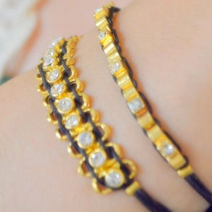 May include: Two gold-toned bracelets with clear, round gemstones and black cord. The bracelets have a decorative, scalloped design. The gemstones are set in gold-colored metal. The bracelets are worn on a wrist.