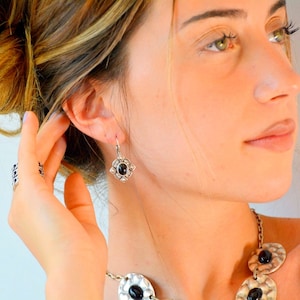 Op de afbeelding: Zilverkleurige oorbellen met een donkere ovale steen in het midden en een bloemmotief. De oorbellen maken deel uit van een sieradenset met een bijpassende ring en ketting met vergelijkbare donkere steenaccenten.