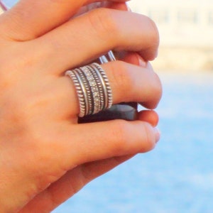 May include: A silver ring with multiple bands, featuring a combination of beaded and smooth textures. Some bands are adorned with small, clear stones. The ring is worn on a finger, showcasing its intricate design and craftsmanship.