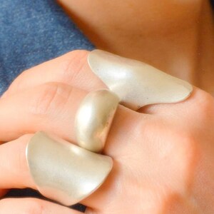 Può includere: Tre anelli argentati di diversi modelli sono mostrati su una mano. Gli anelli hanno superfici lisce e curve. Uno è una semplice fascia, mentre gli altri presentano forme astratte più grandi. Lo sfondo include una giacca di jeans.