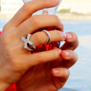 May include: Two silver-toned rings worn on the fingers. One ring features a prominent, flat, cross-shaped design with a matte finish. The second ring is a thin, coiled band with a textured, spiral appearance that wraps around the finger.
