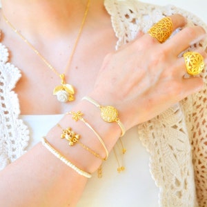 Op de afbeelding: Een verzameling goudkleurige sieraden, waaronder een ketting met een witte rozenhanger, verschillende armbanden en twee sierlijke ringen. De armbanden hebben verschillende ontwerpen en de ringen hebben opengewerkte patronen.