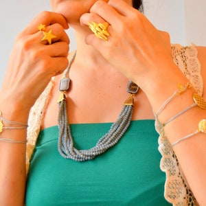 Puede incluir: Conjunto de joyas doradas con un anillo de estrella, un anillo llamativo y múltiples pulseras con dijes decorativos. Un collar de cuentas grises de múltiples hilos con colgantes cuadrados completa el conjunto. Las joyas se muestran en una persona que lleva una camiseta verde.