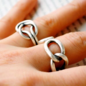 Könnte beinhalten: Nahaufnahme von zwei silberfarbenen Ringen, jeweils mit einem Knotenmuster. Die Ringe werden an Fingern getragen, wobei einer scharf und der andere unscharf ist. Die Ringe haben eine strukturierte, matte Oberfläche.